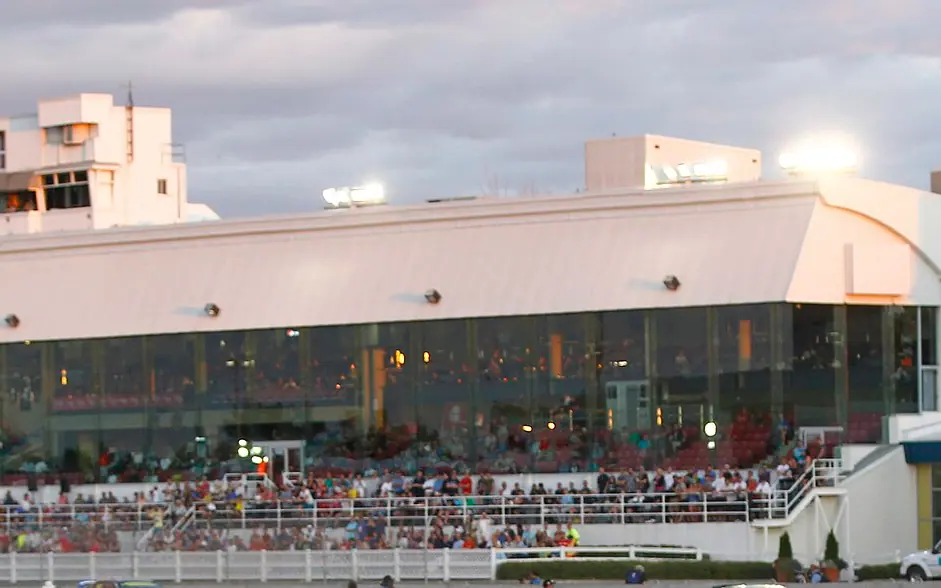 La tribune vitrée de l'Hippodrome de Trois-Rivières, avec des spectateurs le long de la clôture, au crépuscule
